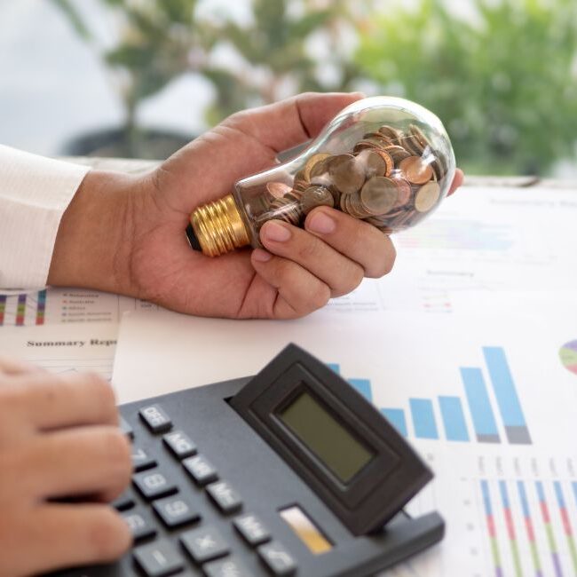 person holding light bulb filled with coins using a calculator